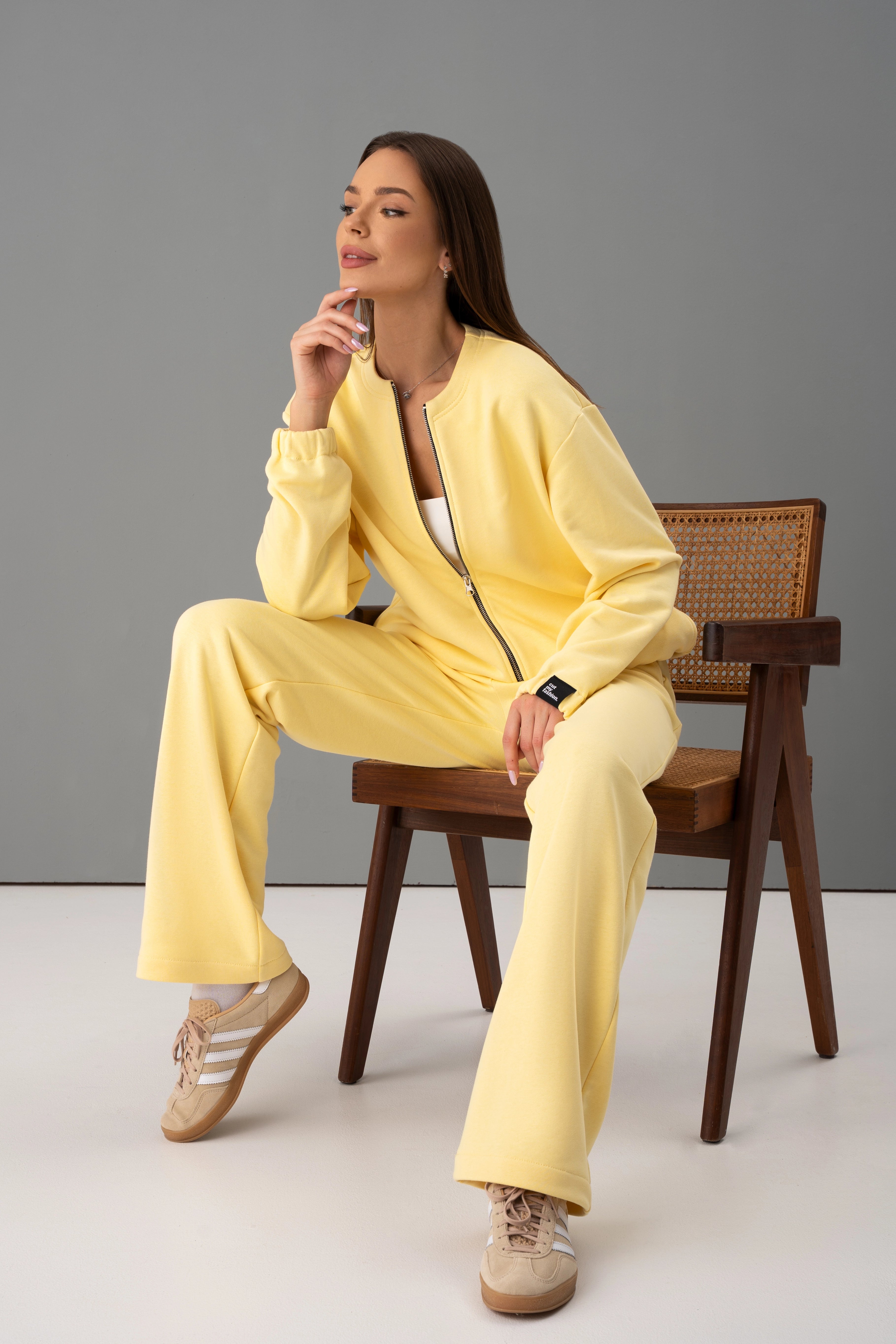 Modelis su geltonais cutmyfashion treningais Mėja CMF547-19 pagamintais Lietuvoje sėdi ant kėdės studijoje, ranka remiasi į smakrą