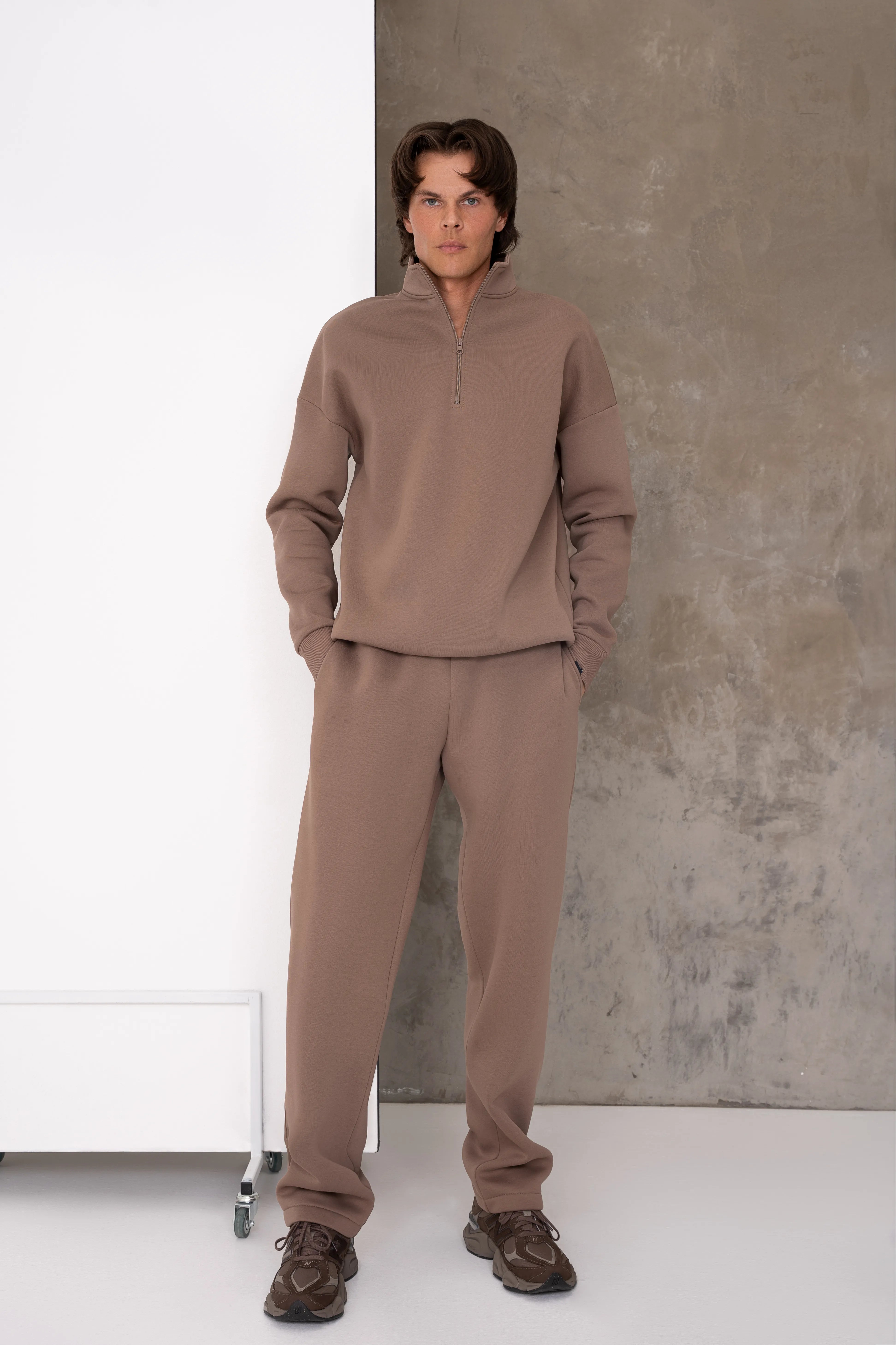 Latte spalvos vyriškas laisvalaikio treningas su aukšta apykakle ir trumpu metaliniu užtrauktuku, nuleistais pečiais, ribo apdaila apačioje ir tiesiomis aukštesnio liemens kelnėmis su raišteliu, pilnas vaizdas iš priekio