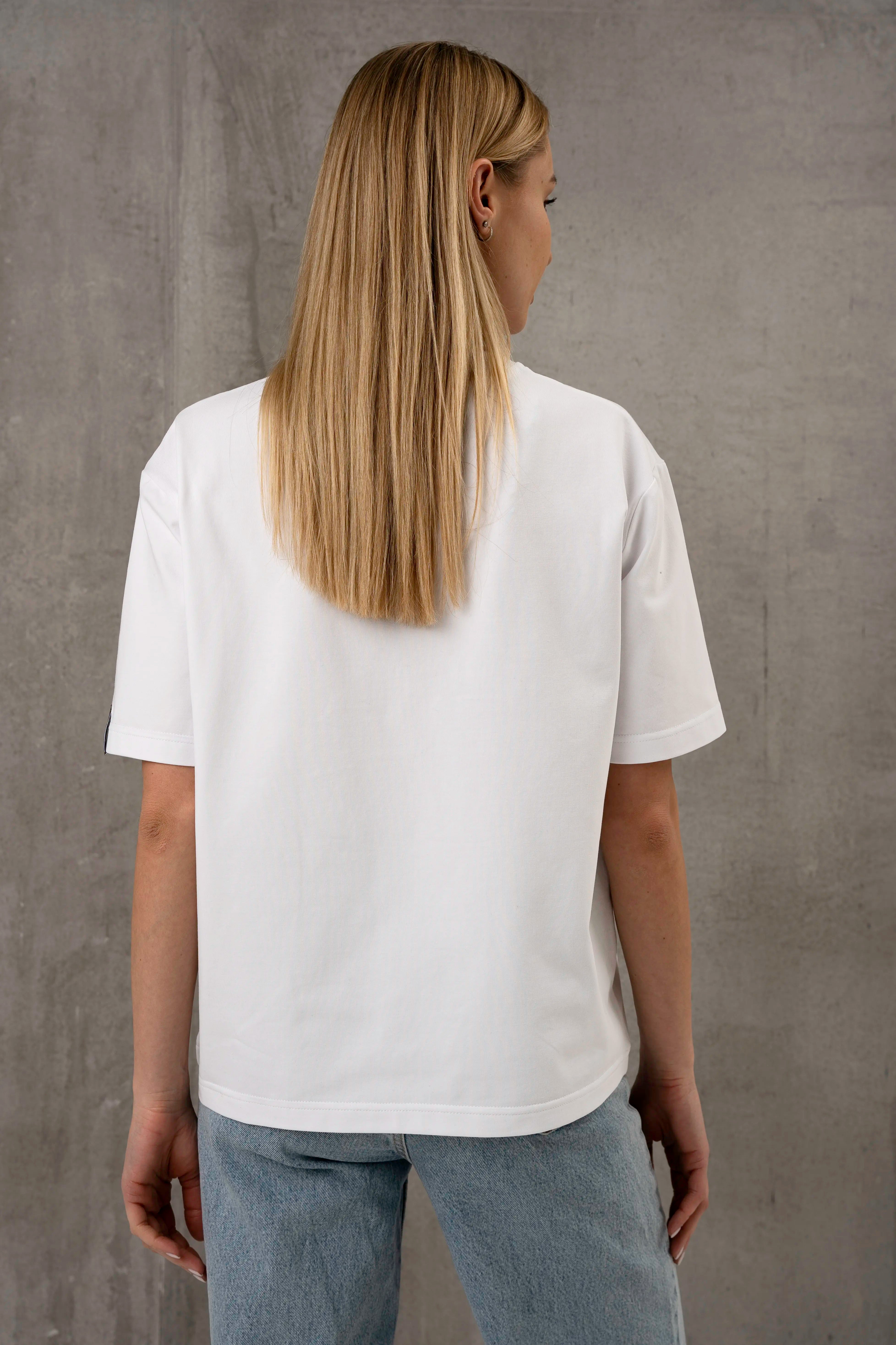 Die Frau trägt ein weißes, übergroßes T-Shirt mit kurzen Ärmeln und helle Jeans; die Aufnahme zeigt sie von hinten vor einem grauen Hintergrund.