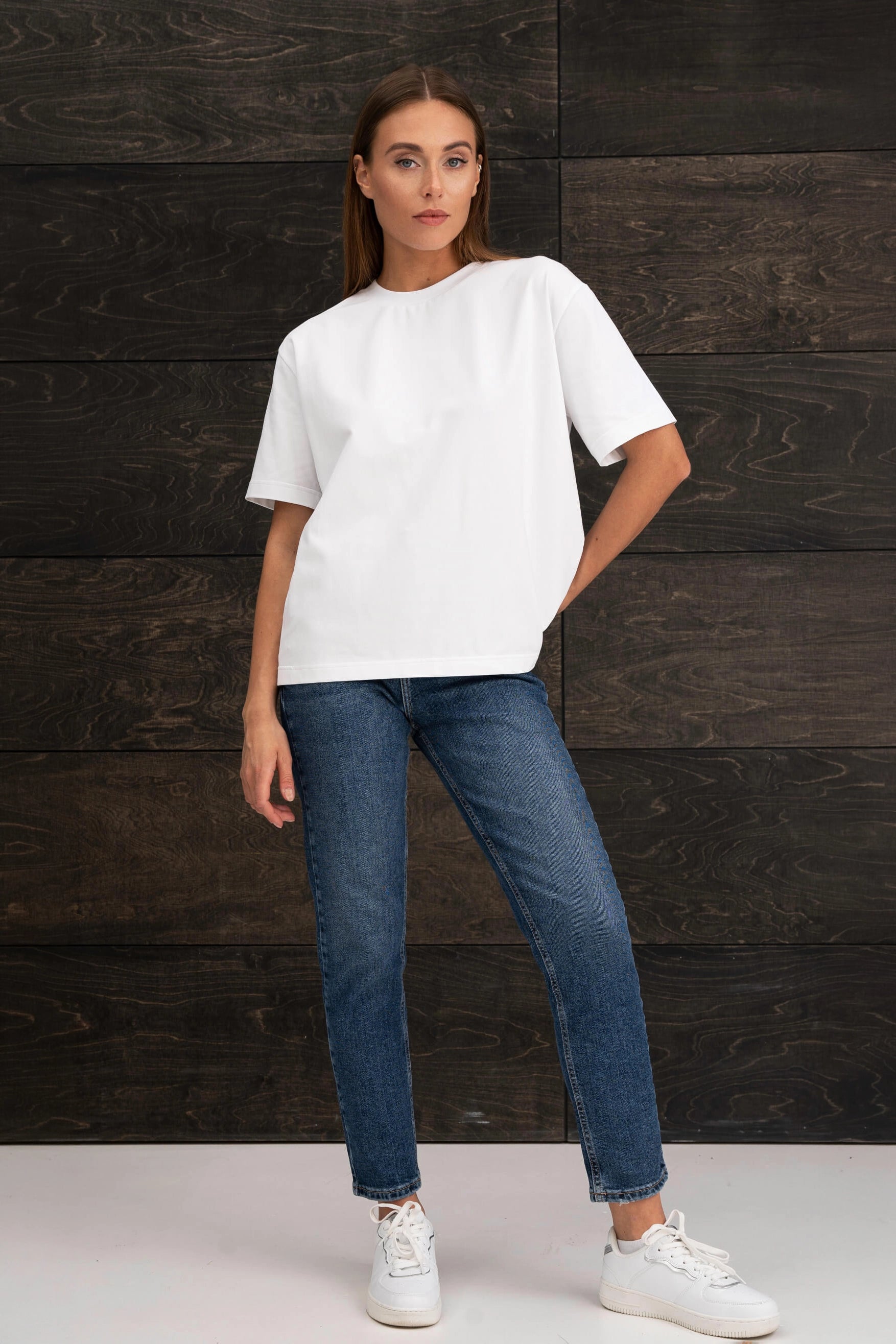 Die Frau trägt ein weißes, übergroßes T-Shirt mit kurzen Ärmeln, eine blaue Jeans und weiße Turnschuhe; sie steht vor einer Wand mit dunklem Holzmuster und hat die Hand in der Tasche.