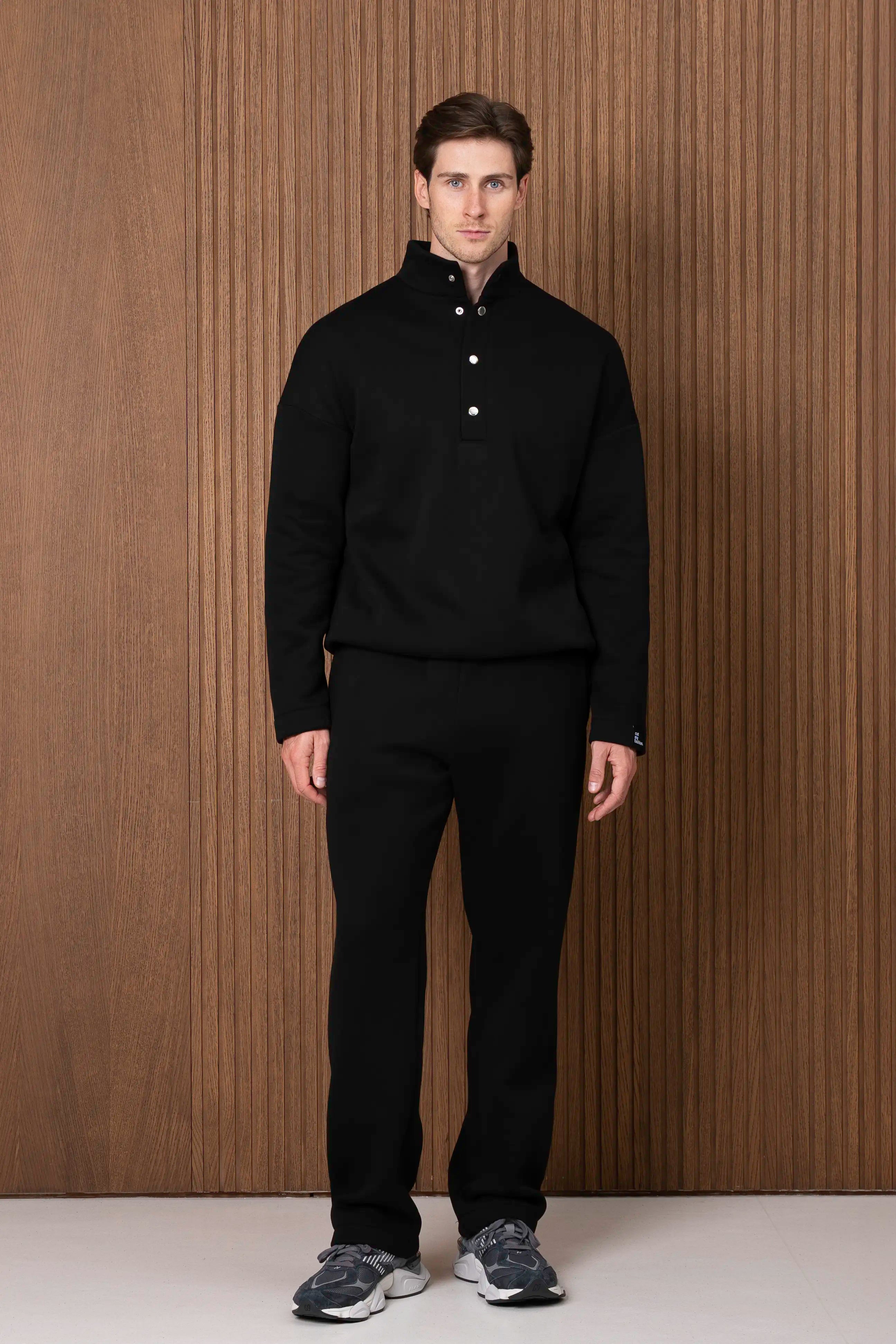Juodas vyriškas laisvalaikio kostiumas trijų ketvirčių kampu, matomas laisvas džemperio siluetas ir tiesios kelnės.
