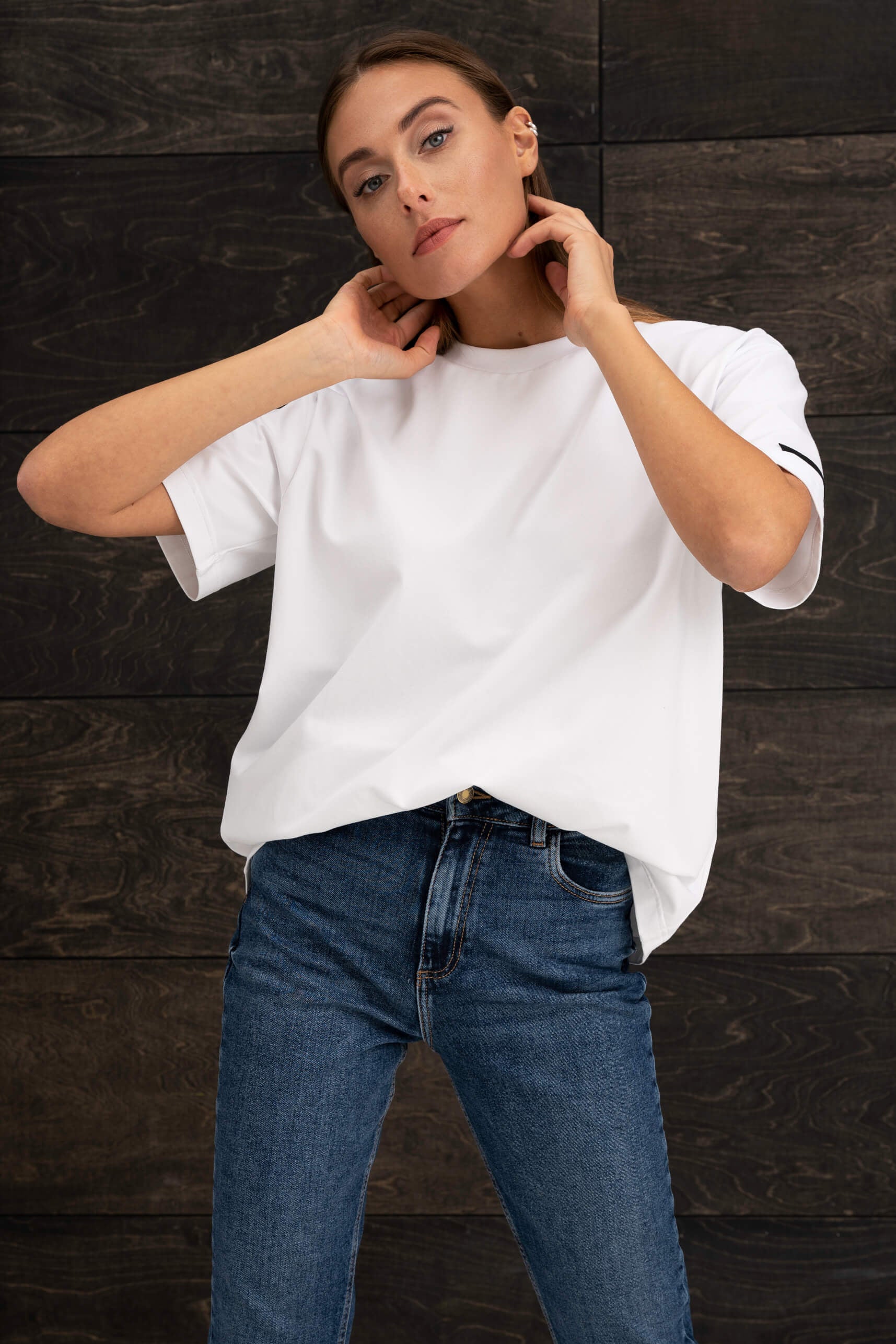 Die Frau trägt ein weißes Oversize-T-Shirt mit kurzen Ärmeln und eine blaue Jeans und steht vor einer Wand mit dunklem Holzmuster.