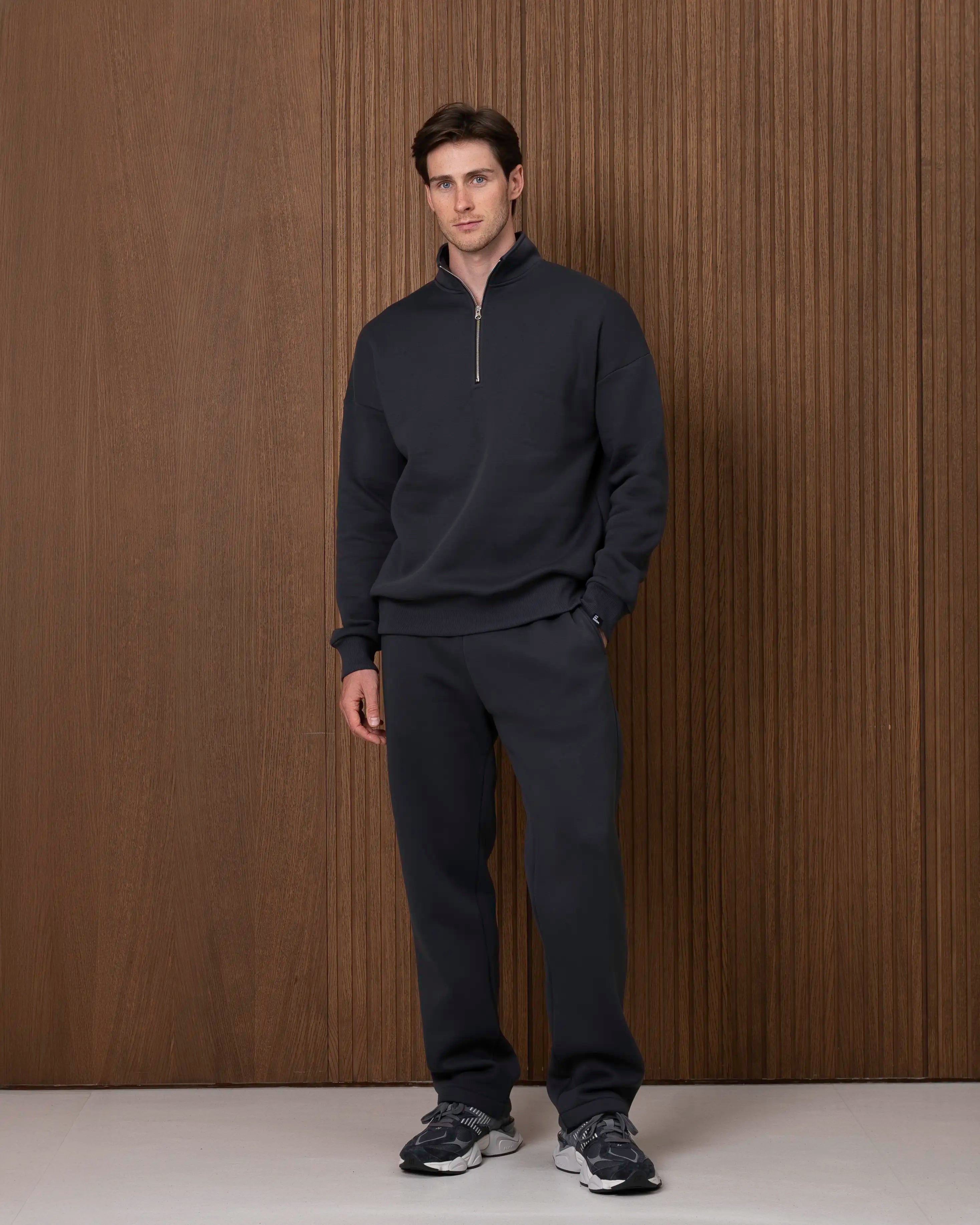Vyras stovi visu ūgiu vilkėdamas tamsiai pilką „Vilius“ laisvalaikio treningą – laisvesnio kirpimo džemperį su aukšta apykakle ir tiesias kelnes.