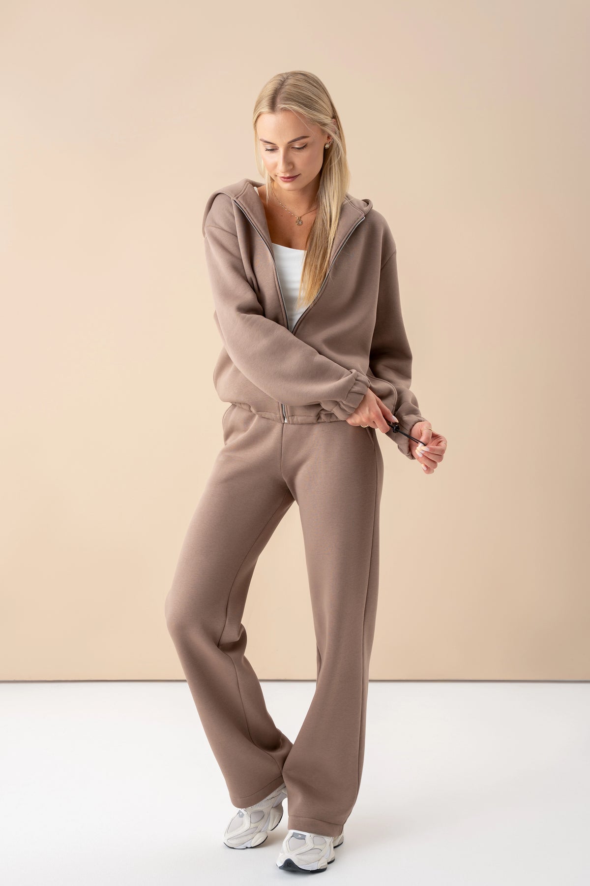 Das Model trägt einen lattefarbenen Trainingsanzug mit Reißverschluss, steht im Studio und schaut nach unten, während sie die Weite des Pullovers anpasst