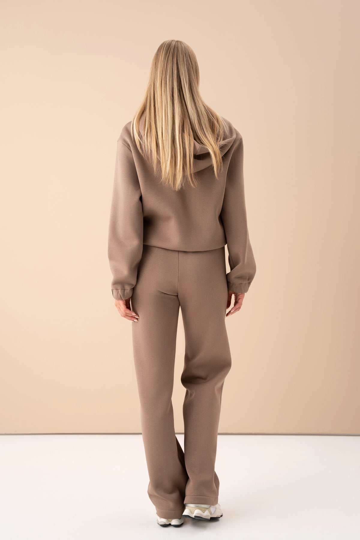 Eine Frau in lattefarbenen Cutmyfashion-Trainingshosen steht mit dem Rücken zur Kamera und posiert vor einem hellen Studiohintergrund
