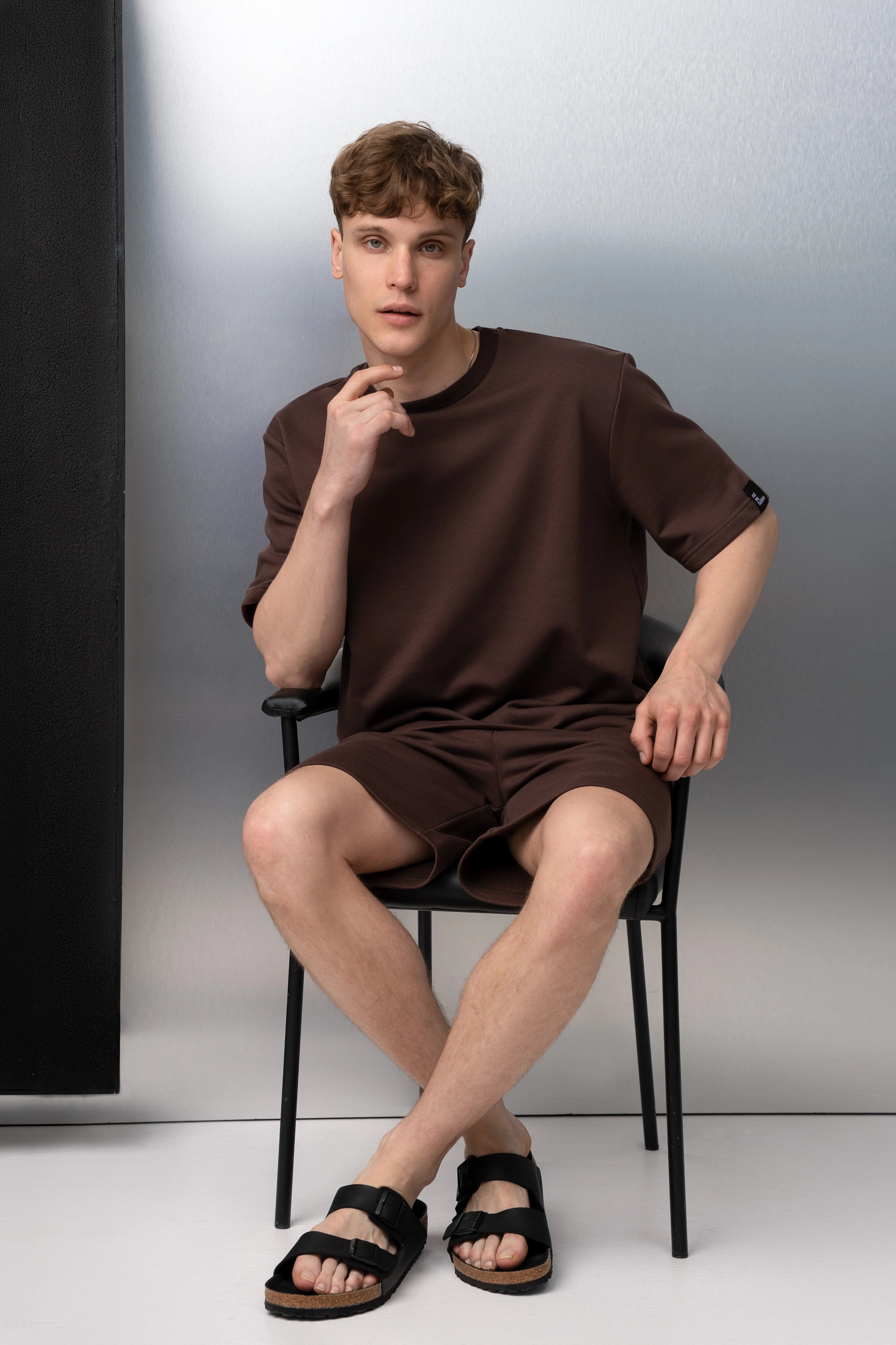 Ein Mann sitzt in einem braunen Anzug aus flauschlosem Stoff, dazu trägt er ein Oversize-T-Shirt und Shorts.