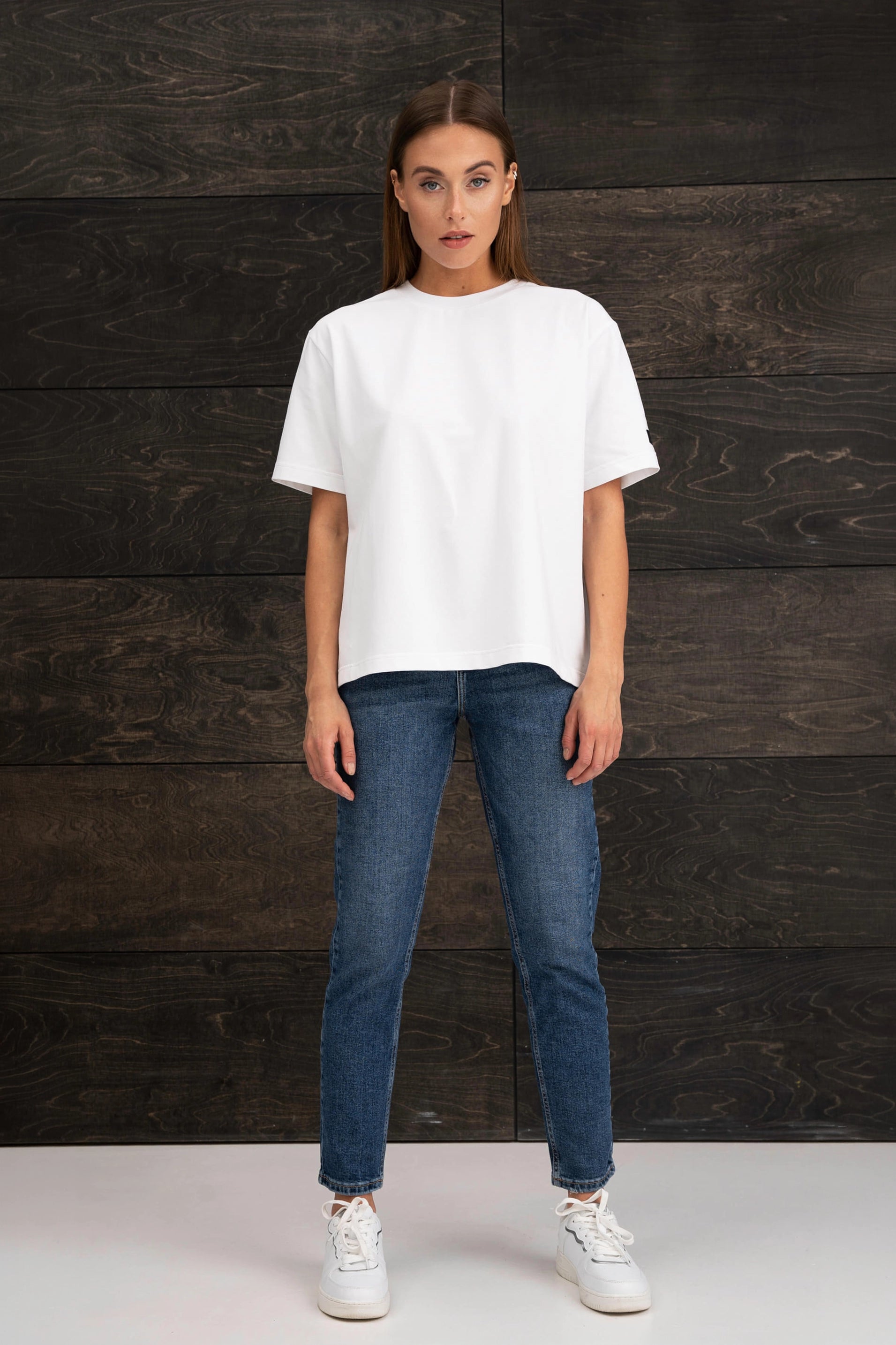 Die Frau trägt ein weißes, übergroßes T-Shirt mit kurzen Ärmeln, eine blaue Jeans und weiße Turnschuhe und steht vor einer Wand mit dunklem Holzmuster.
