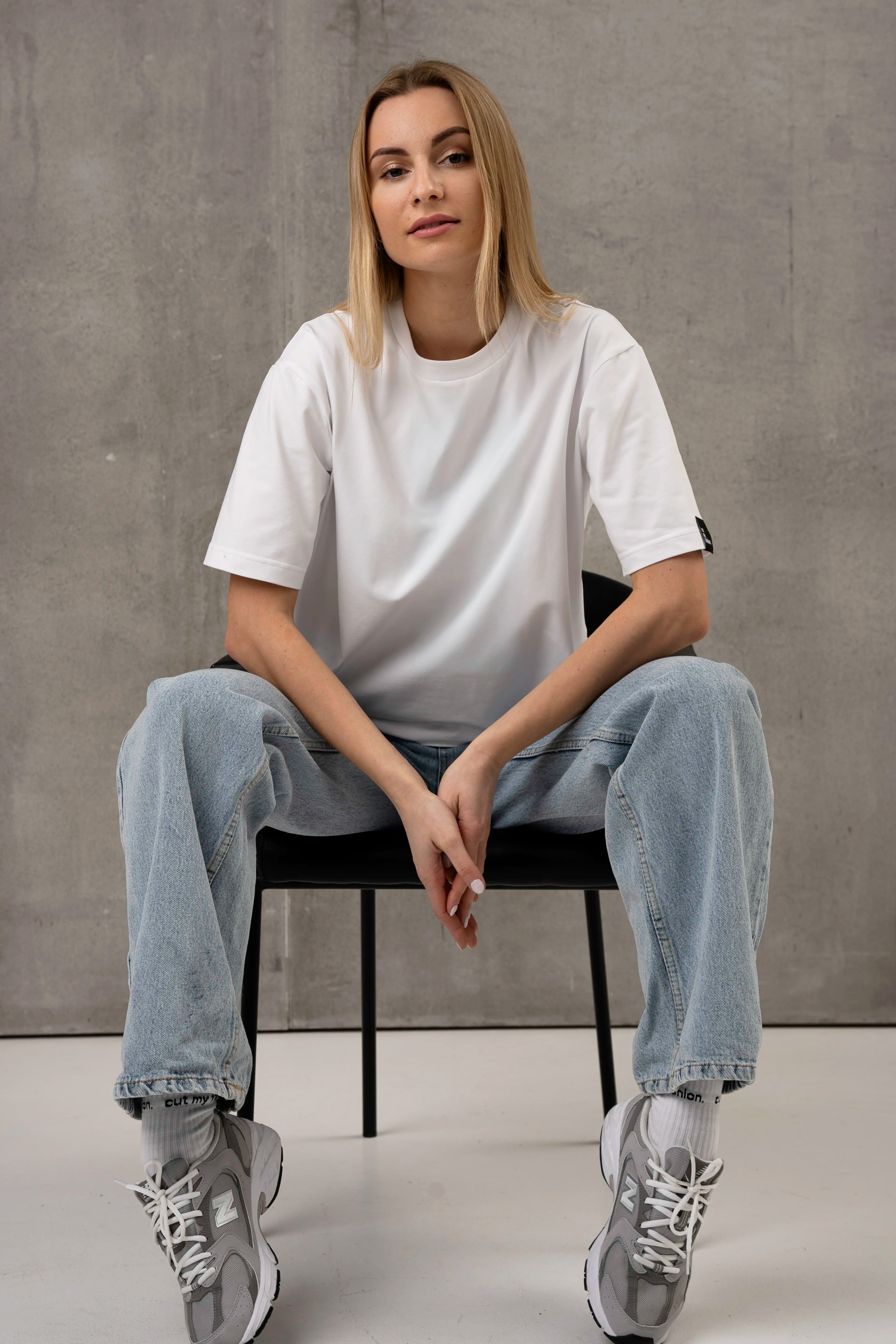 Die Frau trägt ein weißes, übergroßes T-Shirt mit kurzen Ärmeln, helle Jeans und graue Turnschuhe und sitzt auf einem schwarzen Stuhl vor einem grauen Hintergrund.