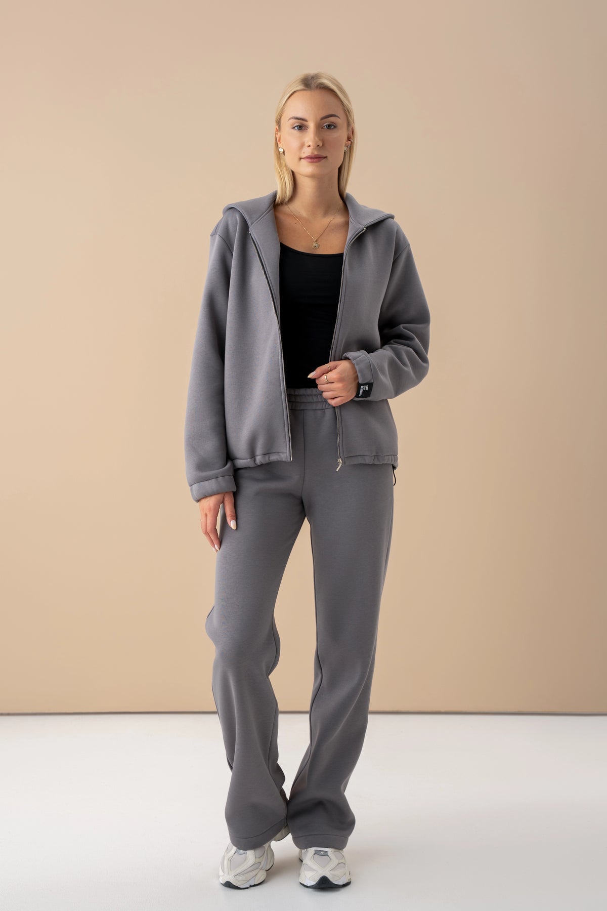 Die Frau trägt einen grauen Trainingsanzug von Cutmyfashion mit einem schwarzen Oberteil; der Pullover ist aufgeknöpft; Studio-Umgebung