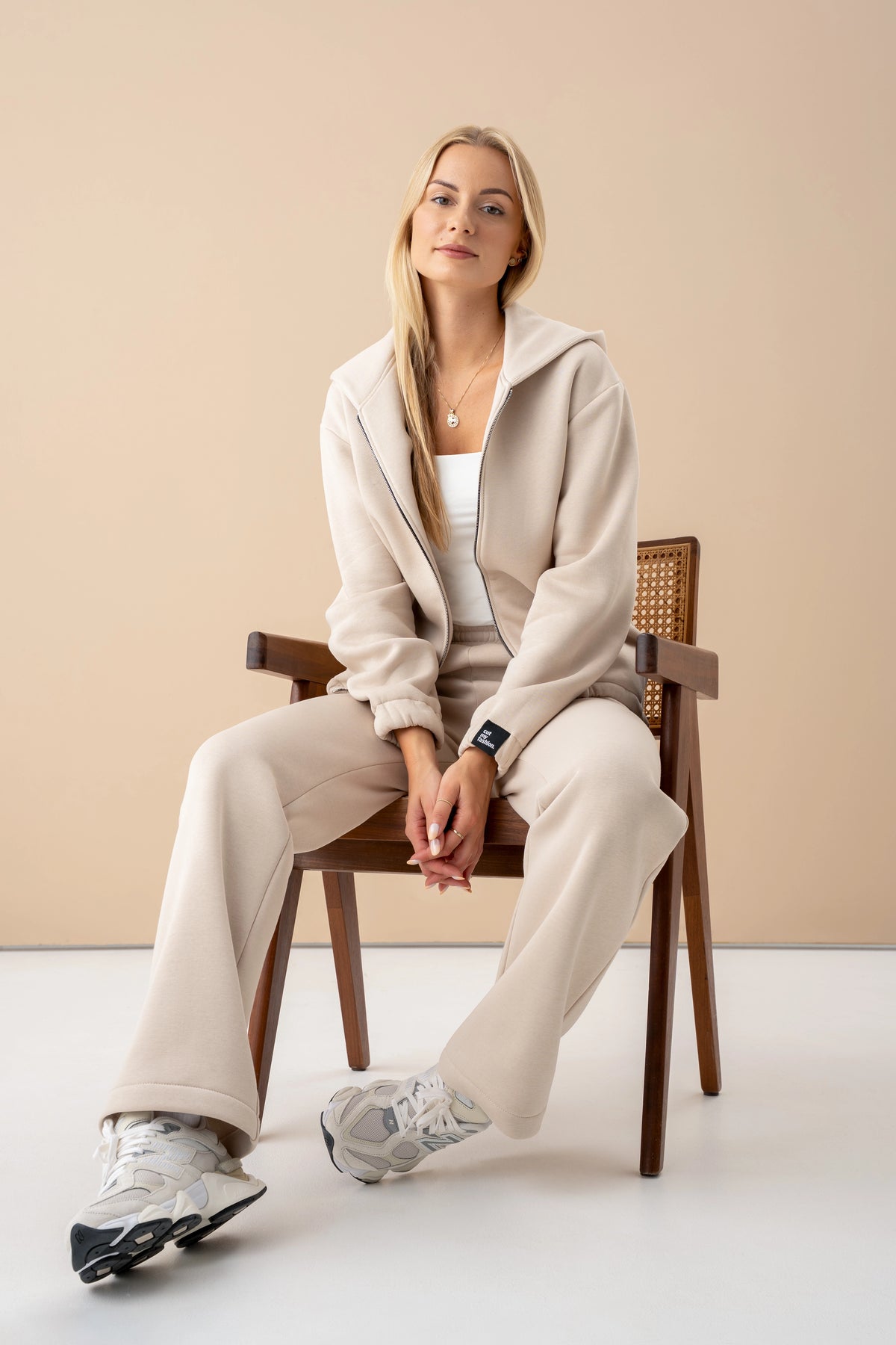 Eine Frau in einer Trainingsanzug von Cutmyfashion sitzt auf einem Stuhl, trägt einen Pullover mit Reißverschluss und schaut in die Kamera