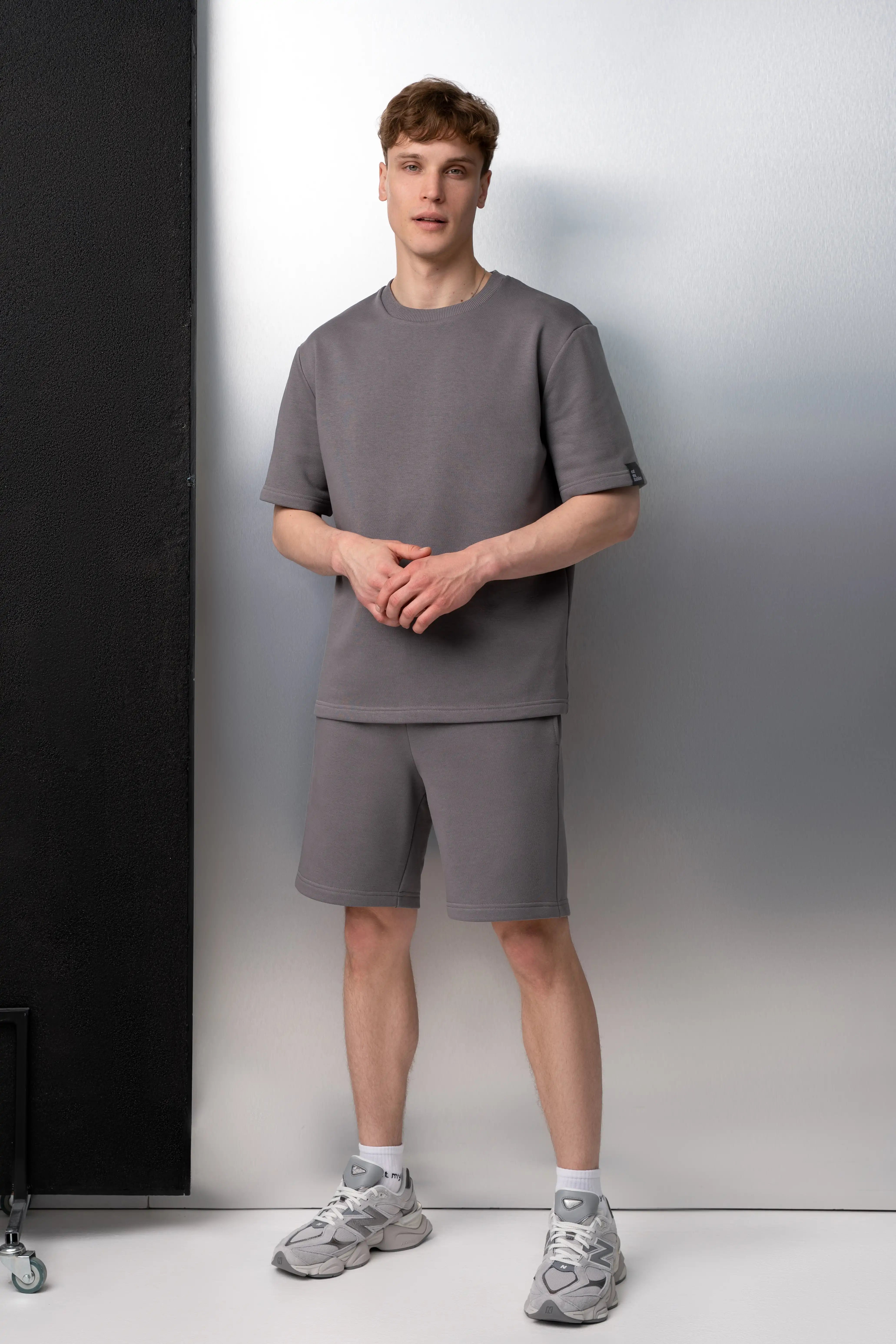 Der Mann trägt einen grauen Freizeitanzug aus flauschfreiem Stoff: ein Oversize-T-Shirt mit kurzen Ärmeln und längere Shorts mit Taschen; die Kombination wurde im Studio fotografiert.