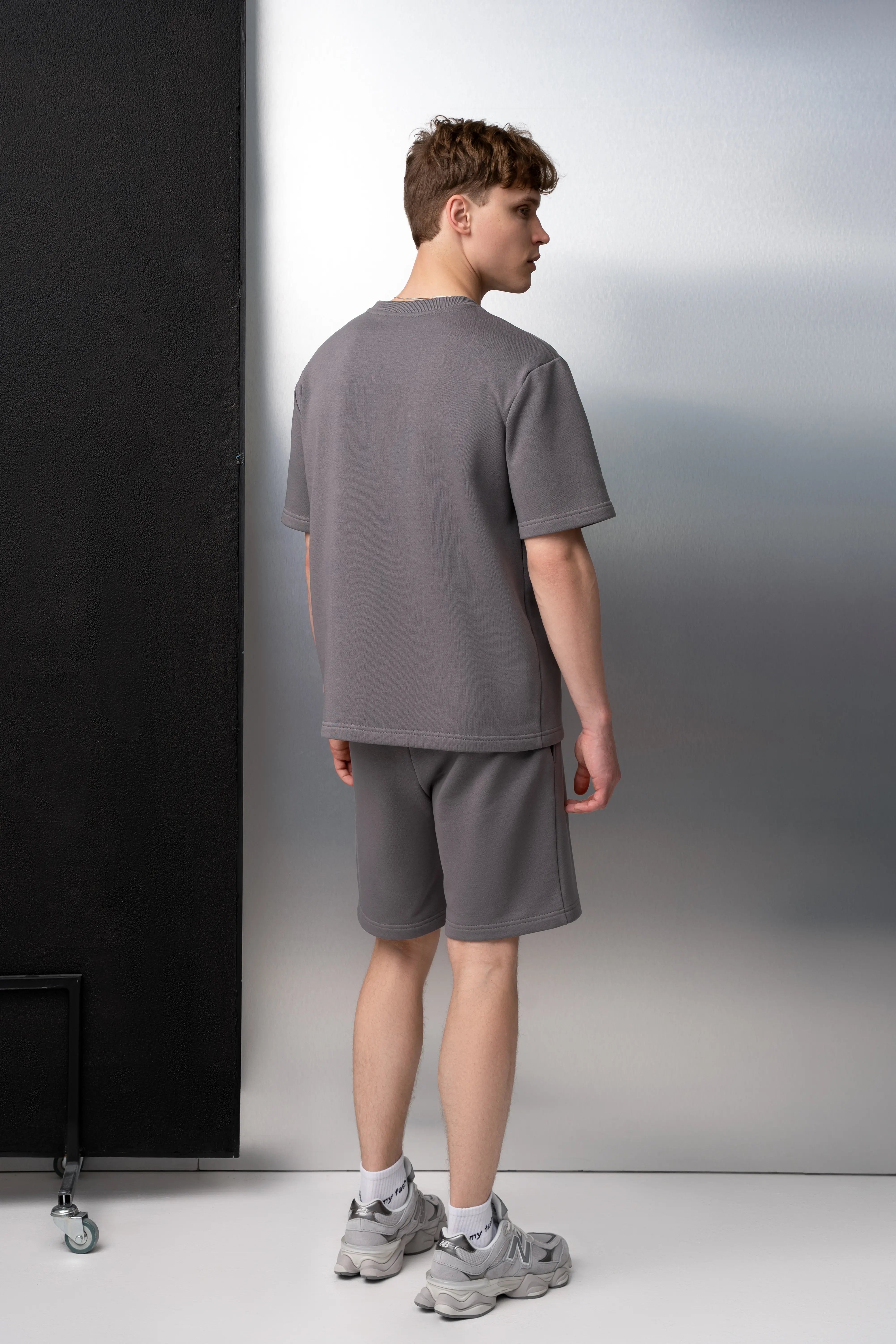 Ein Mann trägt einen grauen Freizeitanzug aus flauschfreiem Stoff: ein Oversize-T-Shirt mit kurzen Ärmeln und längere Shorts mit Taschen; er ist im Studio von hinten zu sehen.