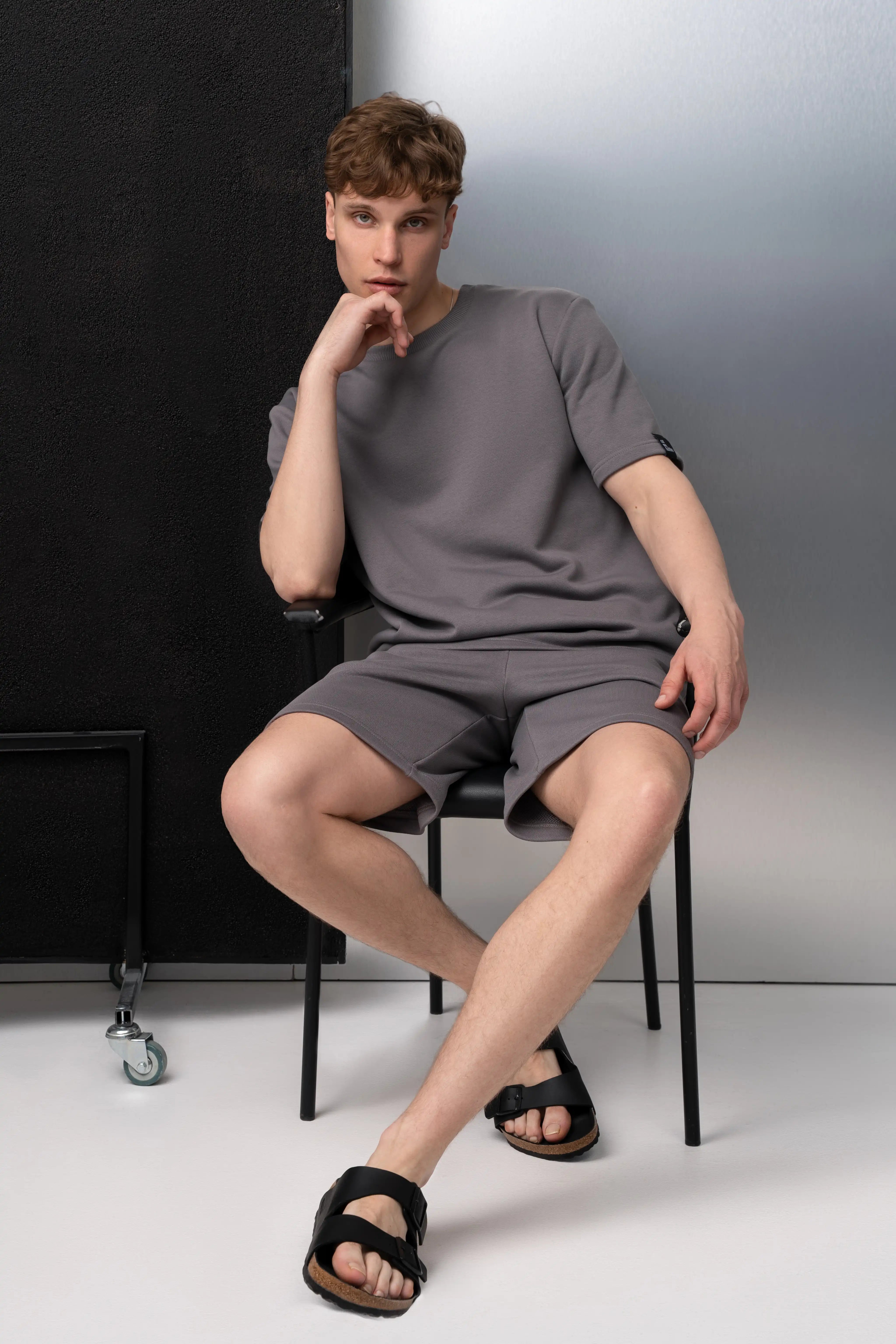 Ein Mann trägt einen grauen Freizeitanzug aus flauschfreiem Stoff: ein Oversize-T-Shirt mit kurzen Ärmeln und längere Shorts mit Taschen; er sitzt auf einem Stuhl im Studio.