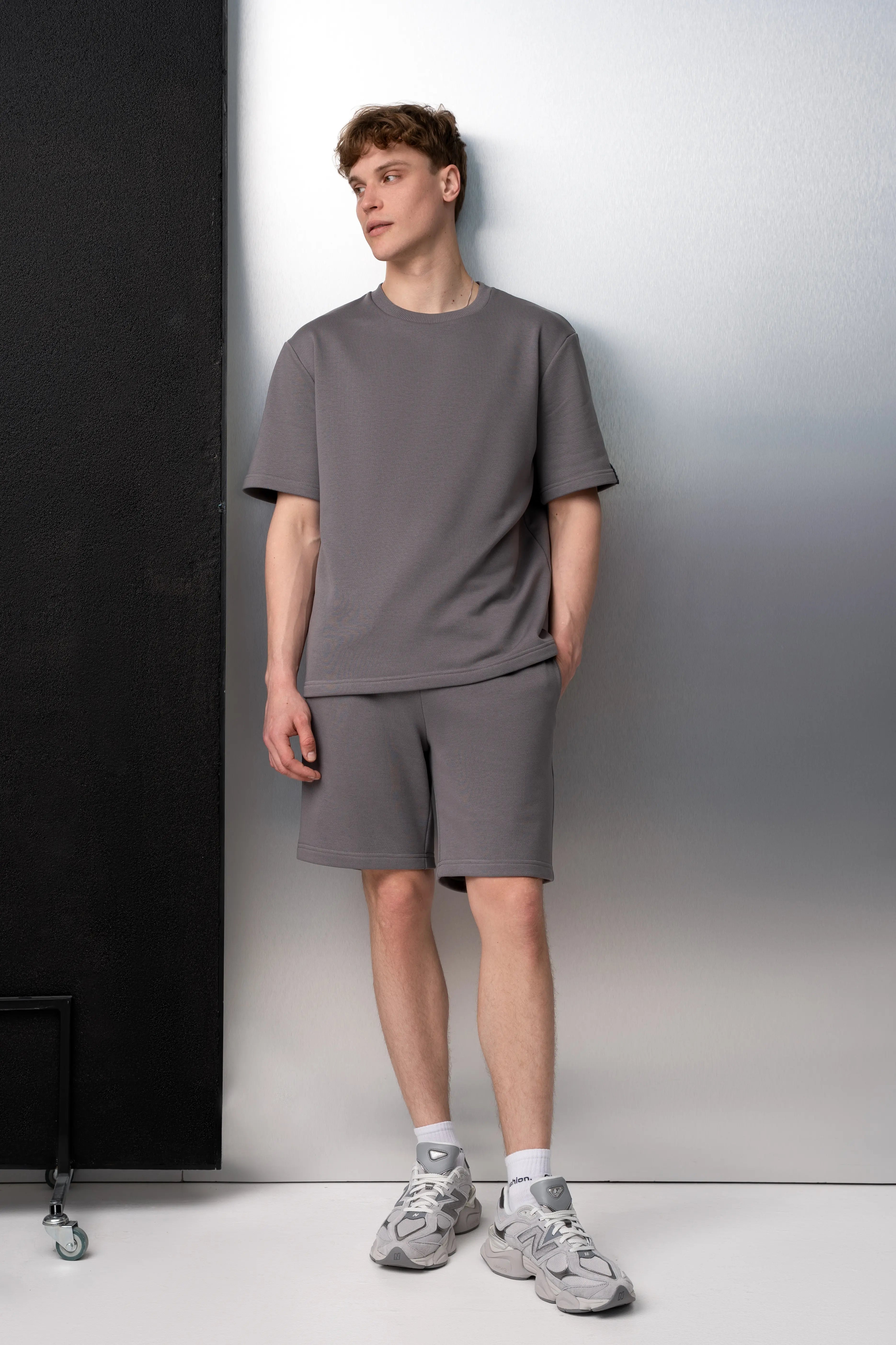 Ein Mann trägt einen grauen Freizeitanzug aus flauschfreiem Stoff: ein Oversize-T-Shirt mit kurzen Ärmeln und längere Shorts mit Taschen; er steht aufrecht im Studio.